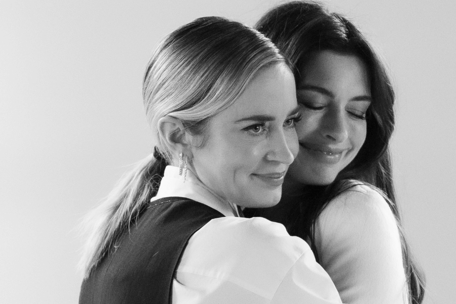 
This black-and-white image captures a tender moment between two women embracing. One has her hair styled back in a sleek ponytail and wears a collared shirt with a vest, smiling softly. The other has long hair and closes her eyes with a content smile, resting her head on the other’s shoulder. The minimalist background enhances the emotional warmth of their connection. 