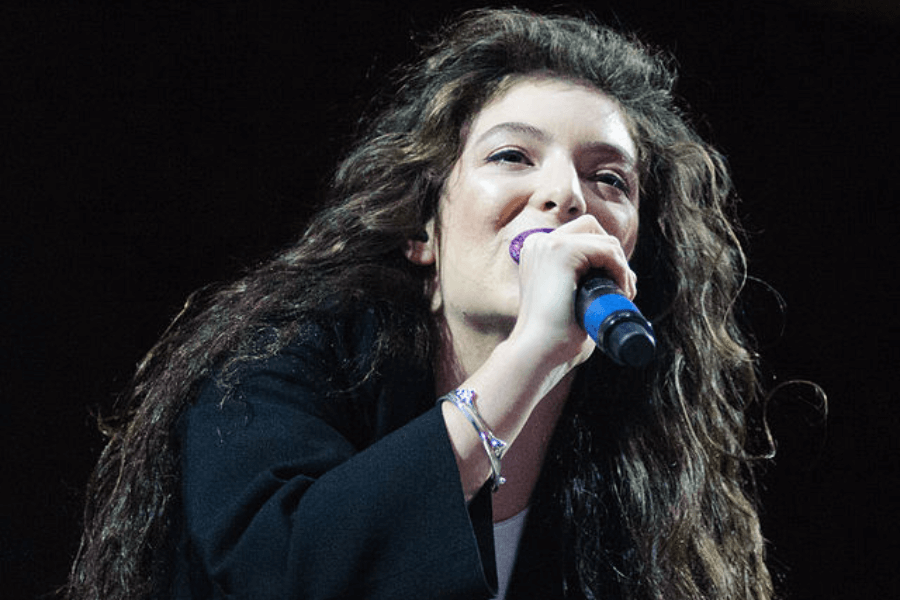 This image shows a performer with long, wavy hair holding a microphone with a blue handle, singing or speaking on stage. They are dressed in dark clothing and wearing a bracelet, with a dimly lit background suggesting a concert or live event.