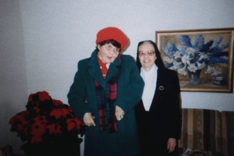 A color photograph of two elderly women posing indoors. One woman is wearing a bright red beret, a green coat, and a plaid scarf, smiling warmly, while the other is dressed in a black outfit with a white collar and glasses. The room is decorated with poinsettias and a floral painting, adding a festive and cozy atmosphere.