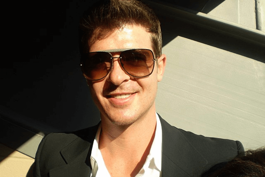 This image features a person wearing aviator-style sunglasses and a dark suit jacket over a white shirt, smiling warmly in a brightly lit setting. Shadows from the surrounding structure add contrast, suggesting the photo was taken outdoors or near a window.