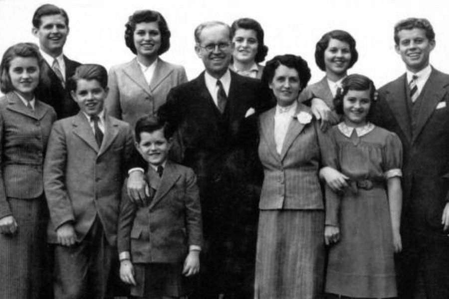 A black-and-white group portrait of a family, featuring parents and several children of various ages. Everyone is dressed in formal attire, with the men in suits and ties and the women in dresses or tailored jackets. The family stands closely together, smiling warmly, capturing a moment of unity and pride.