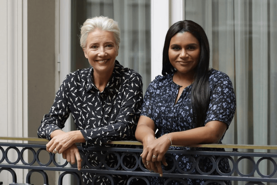 
This image features two women leaning casually on a balcony railing, smiling warmly at the camera. One has short, silver hair and wears a patterned black blouse, while the other has long, sleek dark hair and is dressed in a floral navy outfit. The backdrop includes large windows with sheer curtains, suggesting a sophisticated and relaxed setting. 