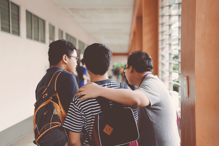 Three students with backpacks walk together in a school hallway. One student has his arm around the shoulder of another, suggesting a moment of friendship and support. The hallway is lined with windows and has an orange-tinted wall. 