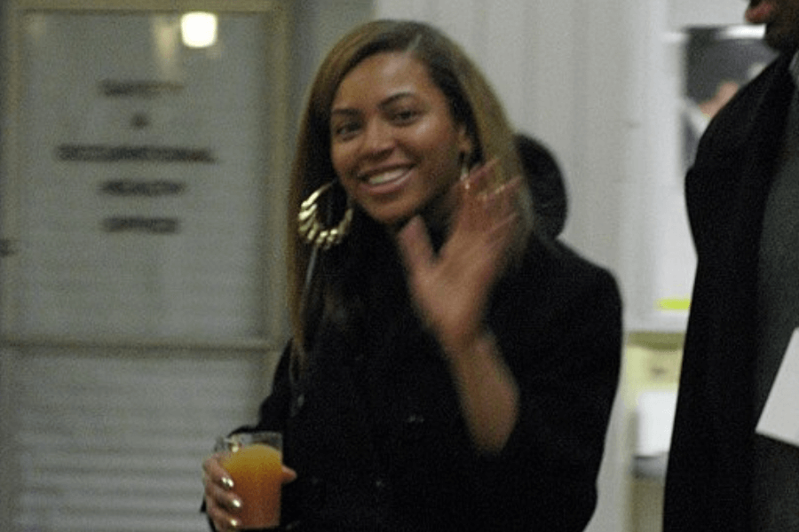This image features a person smiling warmly and waving, dressed in dark attire and holding a glass of orange-colored juice. Large hoop earrings stand out as a notable accessory, and the setting appears to be an indoor space with blurred background details.