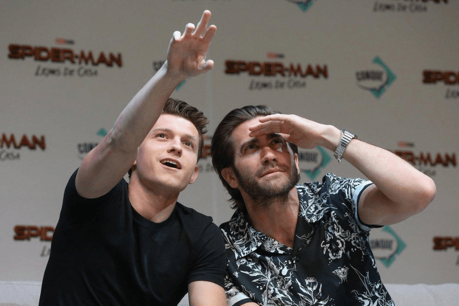 This image shows two men sitting side by side at a press event or panel for "Spider-Man: Lejos de Casa" (Spider-Man: Far From Home). One, wearing a black T-shirt, is gesturing upward with an excited expression, while the other, in a patterned shirt, shades his eyes with his hand as if looking into the distance. The event backdrop features logos and branding for the film, creating a lively and promotional atmosphere.