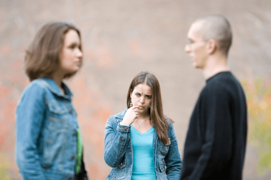 A woman in the background looks upset and confused, with her hand near her face, as she watches two people in the foreground talking. The two people, a woman on the left and a man on the right, are out of focus, while the central figure's expression conveys concern or frustration. 