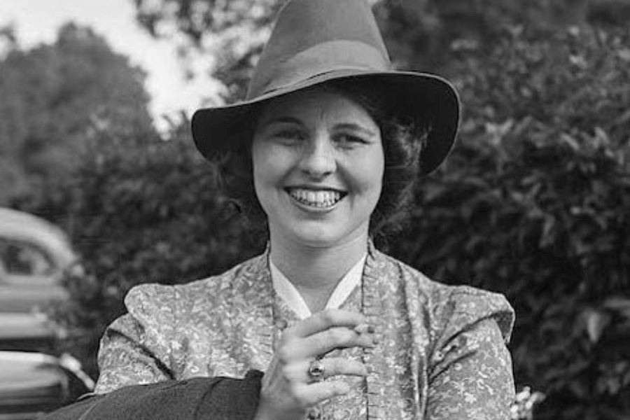 A black-and-white photograph of a woman outdoors, smiling brightly while wearing a wide-brimmed hat and a patterned dress. Her joyful expression and casual pose exude a sense of warmth and confidence, with foliage and part of a car visible in the background, suggesting a relaxed and happy moment.