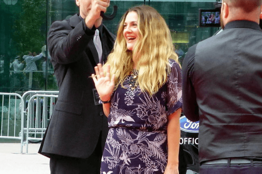 This image features a person with wavy blonde hair wearing a floral patterned outfit, smiling and waving at an outdoor event. Surrounding them are security personnel dressed in formal black attire, with barricades and a glass building in the background, suggesting a red-carpet or public appearance.