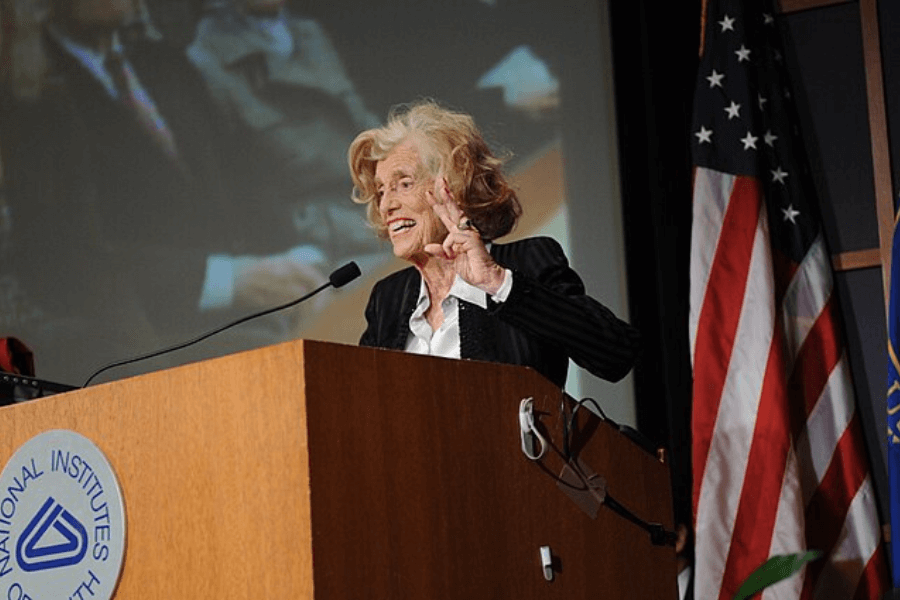 A color photograph of an elderly woman speaking at a podium during an event, gesturing expressively with her hand. She wears a black blazer with white accents and appears passionate and engaged. Behind her are an American flag and a projection screen showing a blurred image of the audience, emphasizing the formal and significant nature of the occasion.