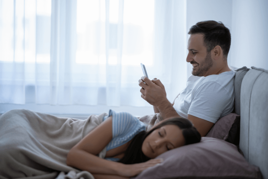 A man with short dark hair, wearing a white t-shirt, sits up in bed smiling while using his smartphone. Beside him, a woman with long dark hair, wearing a striped shirt, sleeps on her side under a blanket, resting on a pillow. The room is softly lit with sheer curtains covering a large window. 