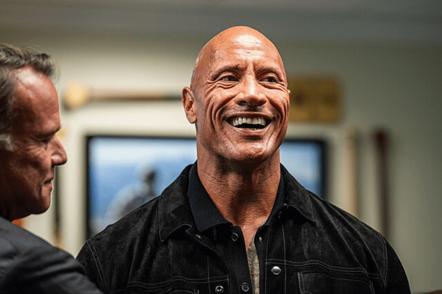 This image features a person smiling widely while wearing a black collared jacket in a brightly lit indoor setting. Another individual is partially visible to the left, engaged in conversation, with blurred background details including framed photos or art.