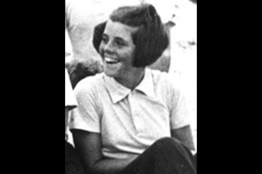 A cropped black-and-white image of a young woman sitting outdoors, laughing and looking to the side. She is dressed in a collared shirt, her short hair styled neatly, exuding a cheerful and carefree demeanor.