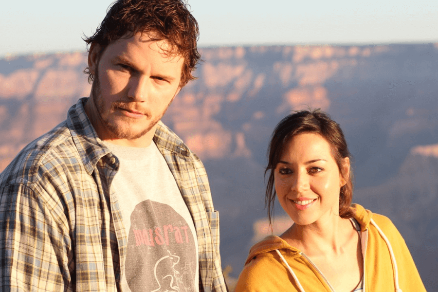 This image features a man and a woman outdoors with a scenic canyon landscape in the background, bathed in warm natural light. The man, wearing a plaid shirt over a graphic T-shirt, has a rugged expression, while the woman, dressed in a yellow hoodie, smiles brightly at the camera. The backdrop suggests a peaceful and picturesque setting, possibly during golden hour. 