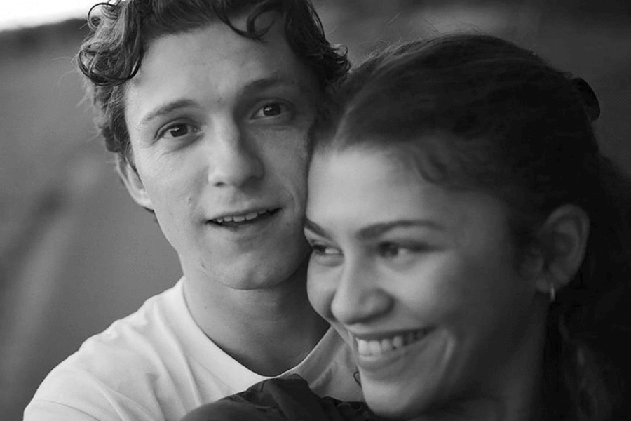 
This black-and-white image captures a candid, intimate moment between a young man and woman. The man, with curly hair and wearing a white shirt, looks directly at the camera with a soft expression, while the woman, with her hair tied back, smiles brightly and leans into him. The blurred background enhances the focus on their close connection and joyful mood.