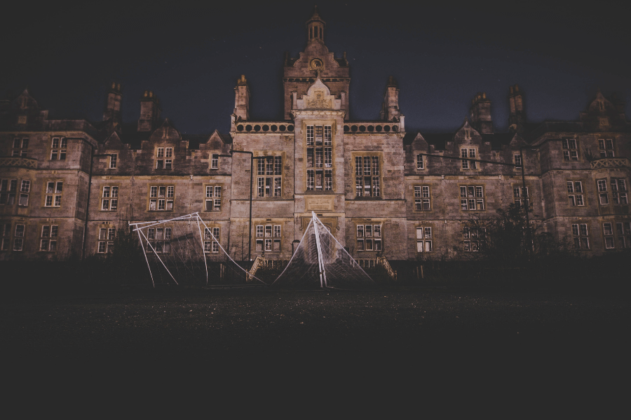  A large, eerie building with gothic architecture stands illuminated against the dark night sky. In the foreground, two broken soccer goals with torn nets are visible on an empty field, adding to the abandoned and haunting atmosphere. 