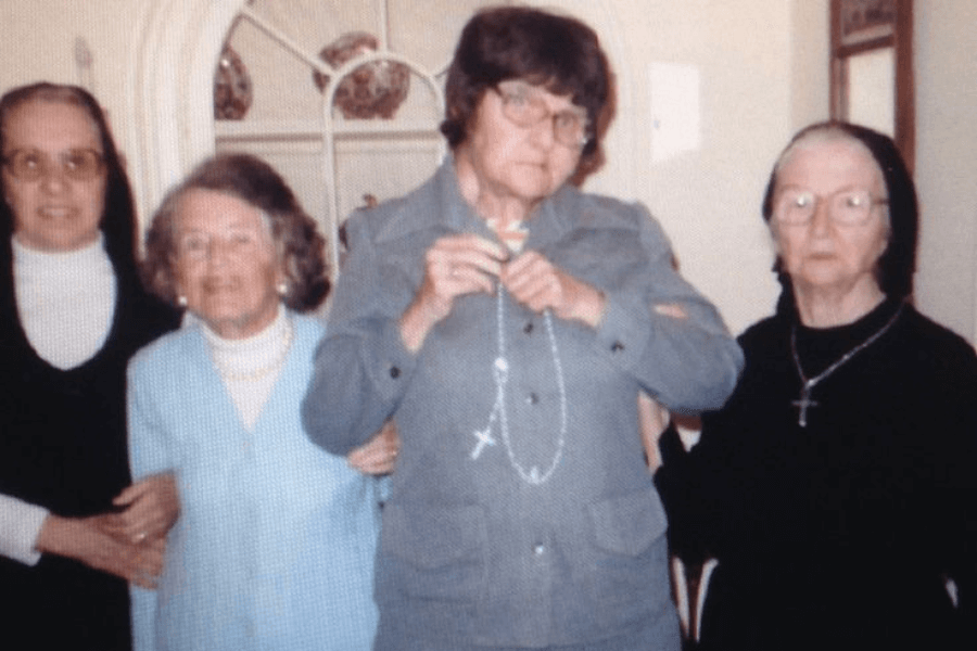 A color photograph of four elderly women standing together indoors, with two dressed in nun habits and wearing crosses. The woman in the center holds a rosary and a small crucifix, emphasizing a religious or spiritual theme. The group is posed in front of a cabinet with decorative items, creating a warm, intimate atmosphere.