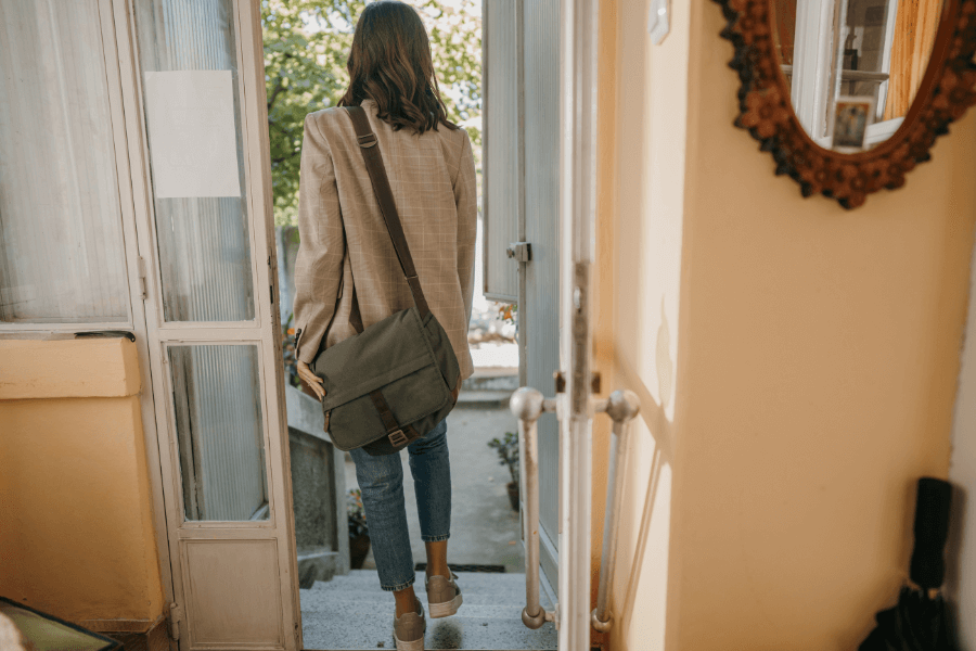 A woman with shoulder-length brown hair, wearing a blazer, jeans, and sneakers, steps outside through an open door. She carries a green messenger bag over her shoulder, and the interior shows a warm, cozy hallway with a decorative mirror on the wall. 