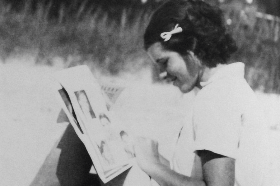 A black-and-white photograph of a young woman seated outdoors, intently reading a magazine or photo album. She has curly hair adorned with a simple bow clip and wears a light blouse, with soft sunlight highlighting her profile. The background features blurred vegetation, suggesting a serene, natural setting.