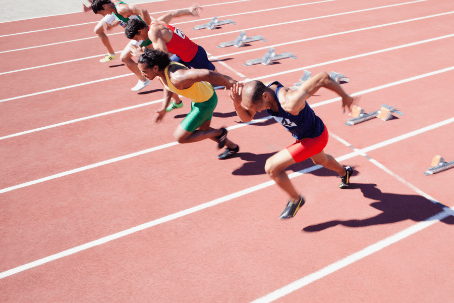 Four male sprinters burst forward at the starting line of a race on a red running track. Their bodies are leaning forward, arms pumping, and shadows stretch across the lanes under bright sunlight. Starting blocks remain behind them. 