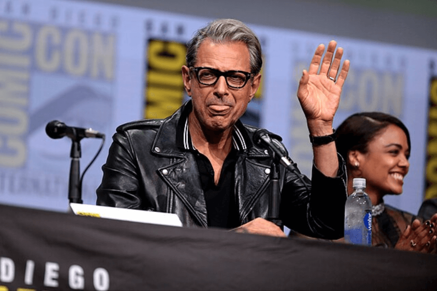 This image shows a person in a leather jacket and glasses speaking at a Comic-Con panel, as indicated by the branded backdrop. They are raising a hand in a friendly gesture, with a microphone and water bottle visible on the table. Another individual seated beside them smiles while looking at the audience.