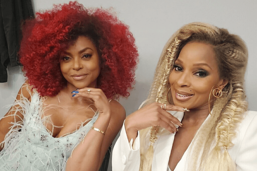 This image shows two women posing with playful expressions. The woman on the left has vibrant red, curly hair and wears a light, feathered dress, resting her chin on her hand with blue-polished nails. The woman on the right has long, styled blonde braids, accessorized with gold hoop earrings, and wears a white outfit, mirroring the pose with her hand near her chin. Both exude confidence and glamour in the photo.