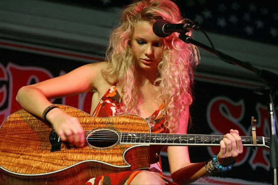 This image features a performer with long, curly hair playing an acoustic guitar on stage under soft lighting. The person appears focused, with a microphone set up nearby, and the background includes partial signage in bold lettering.