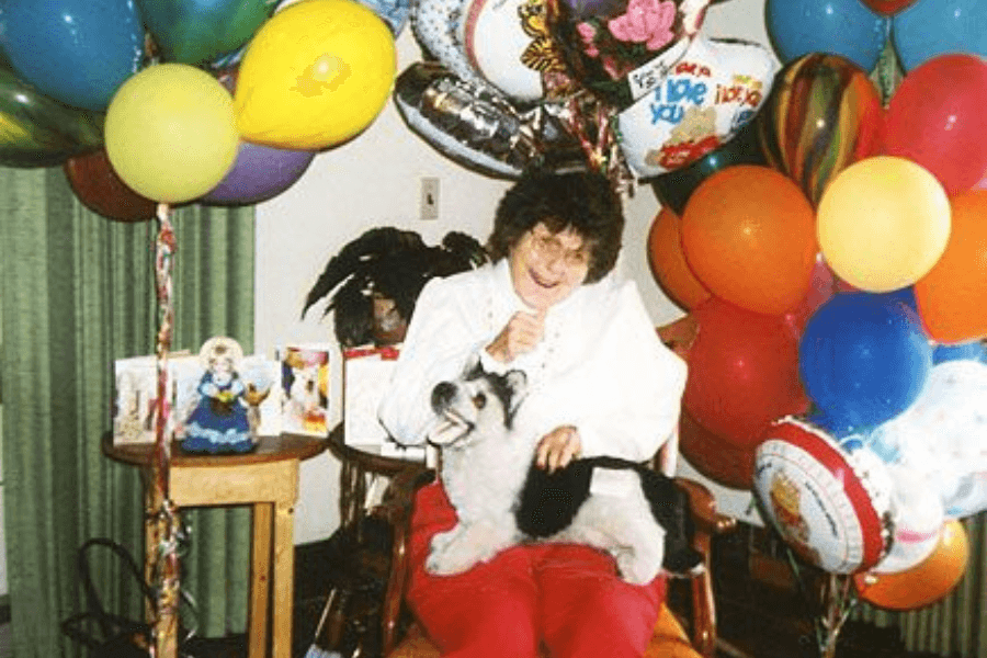 A cheerful scene featuring a woman seated in a chair surrounded by colorful balloons, with a small, fluffy black-and-white dog on her lap. Greeting cards and a festive atmosphere suggest a celebration, possibly a birthday or special occasion. The woman is smiling warmly, adding to the joyful mood.