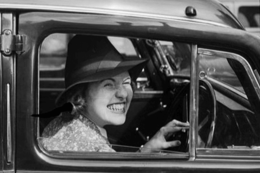 A smiling woman wearing a wide-brimmed hat, seated in the driver’s seat of a vintage car, exudes joy and confidence. The black-and-white image captures the nostalgic charm of an earlier era, showcasing both the woman’s lively expression and the car’s classic design.