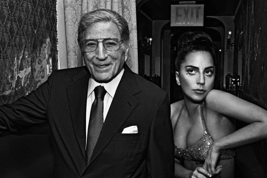 This black-and-white image features an older man in a suit and tie, wearing glasses, standing confidently next to a glamorous woman dressed in a glittering halter-top outfit. The woman has her hair styled in an elegant updo, striking a poised and dramatic expression. The scene exudes a sense of timeless sophistication.