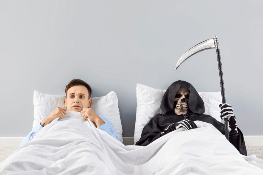 A man with a fearful expression sits in bed under a white blanket, clutching it tightly. Beside him, the Grim Reaper, dressed in a black hooded cloak and holding a large scythe, lies in bed, grinning with a skeletal face. The setting is a minimal bedroom with plain gray walls and white pillows.