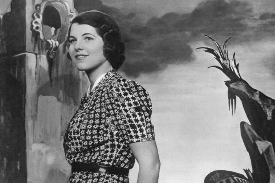 A black-and-white portrait of a woman standing in front of a surreal backdrop featuring abstract architectural elements and dramatic skies. She wears a patterned dress with a fitted waist and a composed smile, her hair styled in soft waves, evoking a classic, vintage aesthetic.