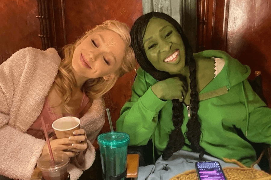 This image depicts two women sitting closely and smiling warmly, suggesting a relaxed, friendly moment. One woman, with blonde hair, wears a soft pink outfit and holds a cup, leaning her head gently. The other woman has green makeup and braided hair, dressed casually in a green hoodie, with a vibrant smile. Drinks and a phone rest nearby, adding to the casual, behind-the-scenes vibe.