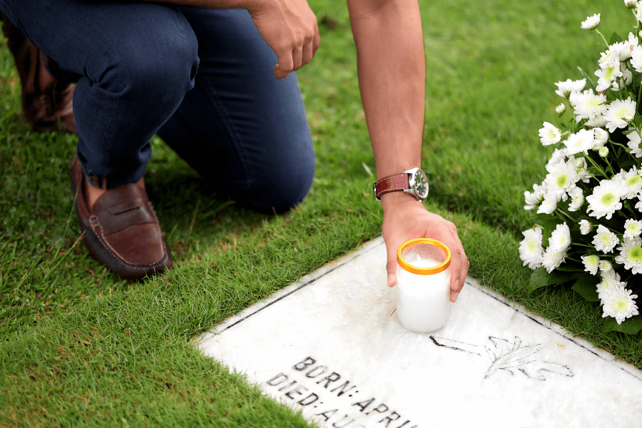 A person kneeling at a gravesite holds a lit candle in a glass container over a marble headstone. The headstone features engraved text and a flower design, surrounded by green grass and a bouquet of white flowers nearby. The person is wearing dark jeans, a watch, and brown shoes. 