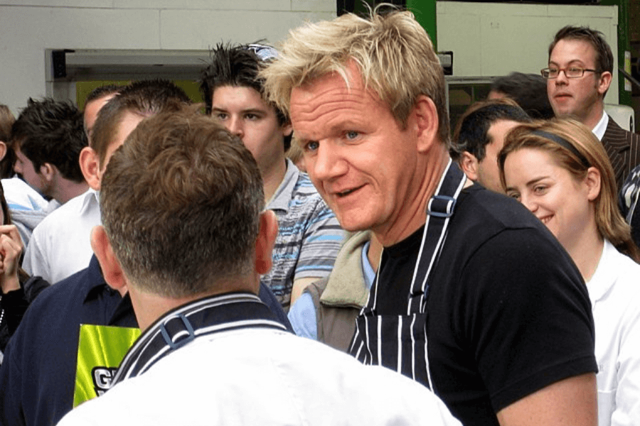 This image shows a person wearing a striped apron and black shirt, engaged in conversation with another individual in a crowded outdoor setting. The crowd includes people of various ages, appearing to participate in or observe an event, possibly culinary-related.