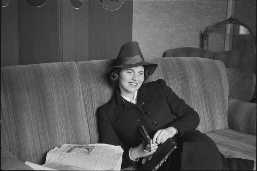 A black-and-white photograph of a woman lounging comfortably on a striped sofa, wearing a tailored coat and a wide-brimmed hat. She is holding a small purse and glancing towards the camera with a relaxed smile, while a folded newspaper rests nearby, capturing a moment of leisure in a vintage setting.