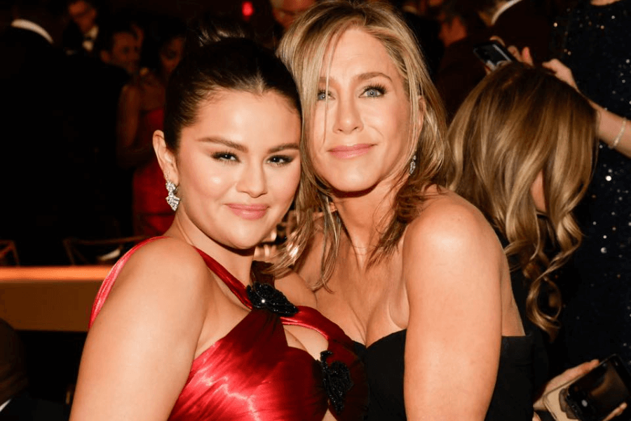 
This image shows two women posing together in an elegant, formal setting, likely at an event or gala. One is wearing a striking red gown with black accents and statement earrings, while the other is in a classic black dress with soft waves in her hair. Both are smiling warmly at the camera, surrounded by a lively crowd in the background. 