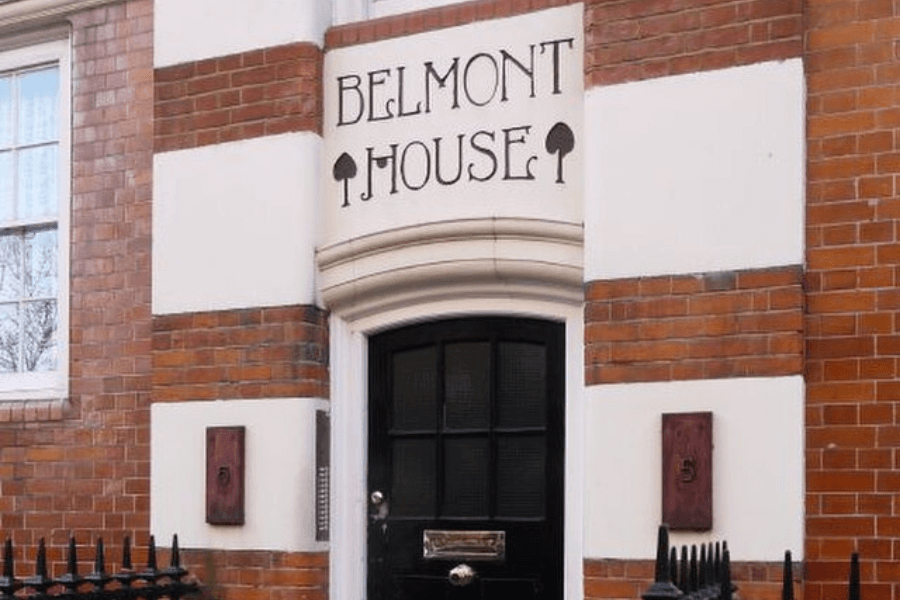 A photograph of the entrance to "Belmont House," as indicated by a decorative sign above the door with stylized spade symbols. The building features red brick and white accents, with a black door and brass mail slot, framed by vintage architectural details and an iron fence in the foreground.