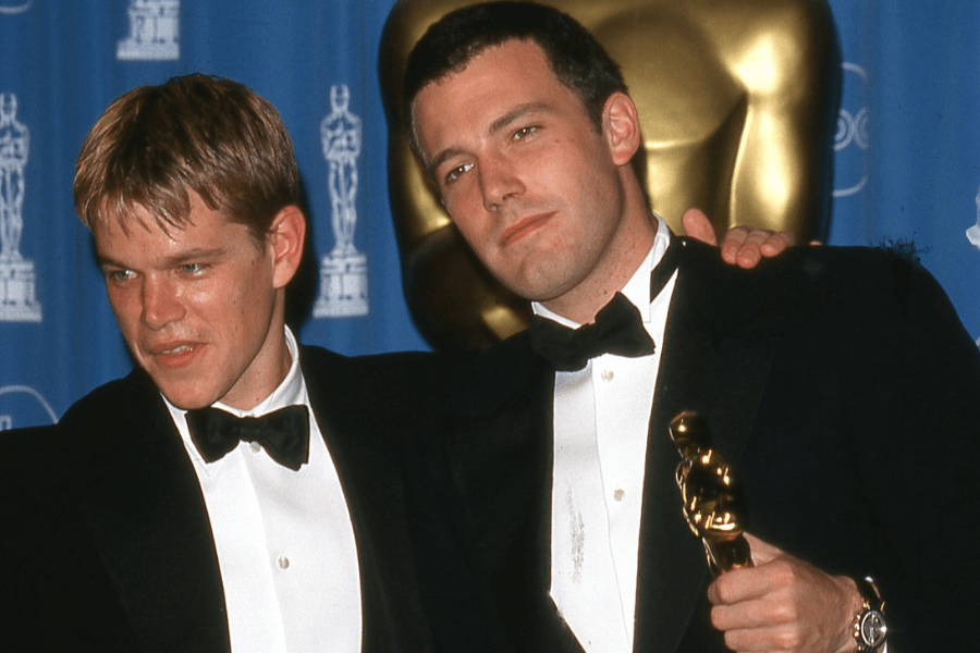 
This image captures two men in tuxedos at an awards ceremony, standing in front of a backdrop featuring Oscar statuette graphics. One man has his arm around the other, smiling slightly, while holding an Oscar trophy in his hand. The atmosphere is celebratory, with a classic formal event vibe. 