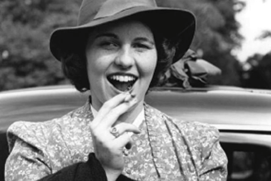 A black-and-white photograph of a woman smiling playfully while holding a small object near her mouth, possibly a cigarette. She is wearing a wide-brimmed hat and a patterned blouse, standing outdoors with a car in the background, exuding a carefree and lively attitude.