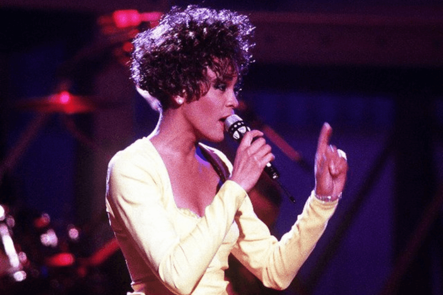 This image shows a performer in a bright yellow outfit holding a microphone while singing on stage, with dramatic lighting and musical equipment in the background. Their expressive gesture, with one hand raised, captures the energy and emotion of the performance.