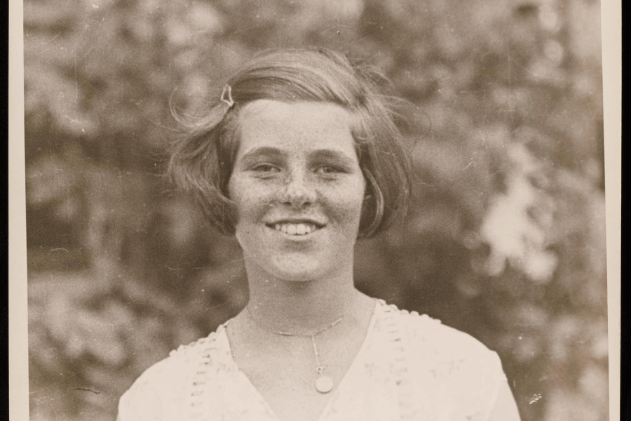 A sepia-toned portrait of a young woman with a short bob haircut, freckles, and a bright smile, wearing a delicate necklace and a light, embroidered blouse. The background is softly blurred, featuring foliage, giving the photo a warm and nostalgic atmosphere.