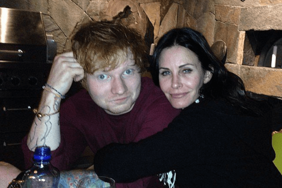 This image shows a man and a woman sitting closely together in a cozy, rustic setting with a stone wall and fireplace in the background. The man, with red hair and casual attire, rests his head on his hand, while the woman leans in, smiling and wrapping her arms around him. The relaxed and friendly atmosphere suggests a casual, personal moment.