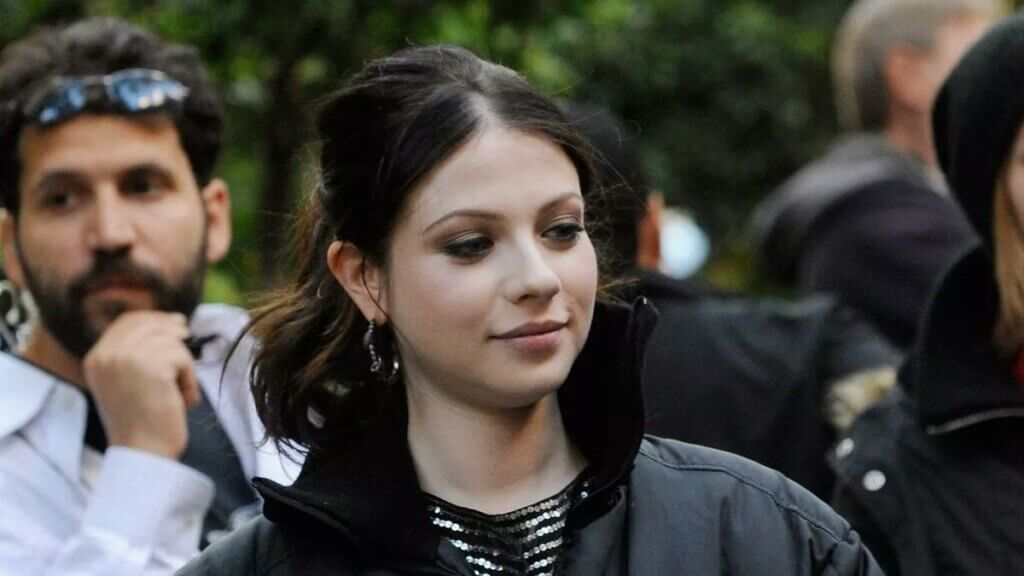 A young woman with dark brown hair styled in a loose updo is seen wearing a black puffer jacket over a sparkly top. She has subtle smoky eye makeup and dangling earrings, with a soft smile on her face. The background is slightly blurred, showing a crowd of people, including a man with sunglasses resting on his head.