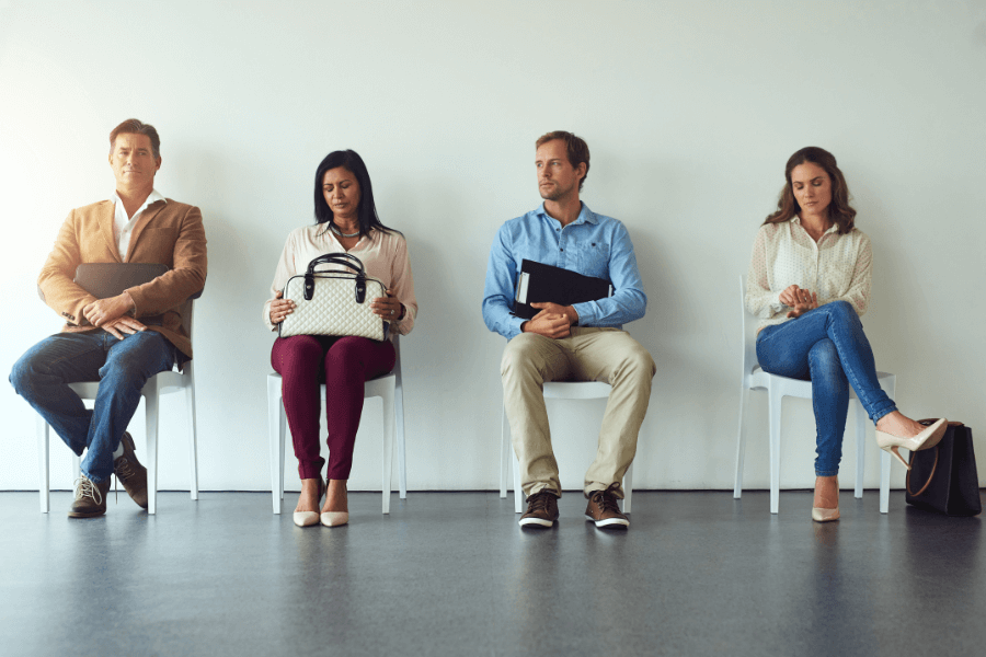 This image shows four people sitting in a row on chairs, each appearing to wait for an interview or meeting. They are dressed in business casual attire, holding items like a handbag, a folder, or a laptop.