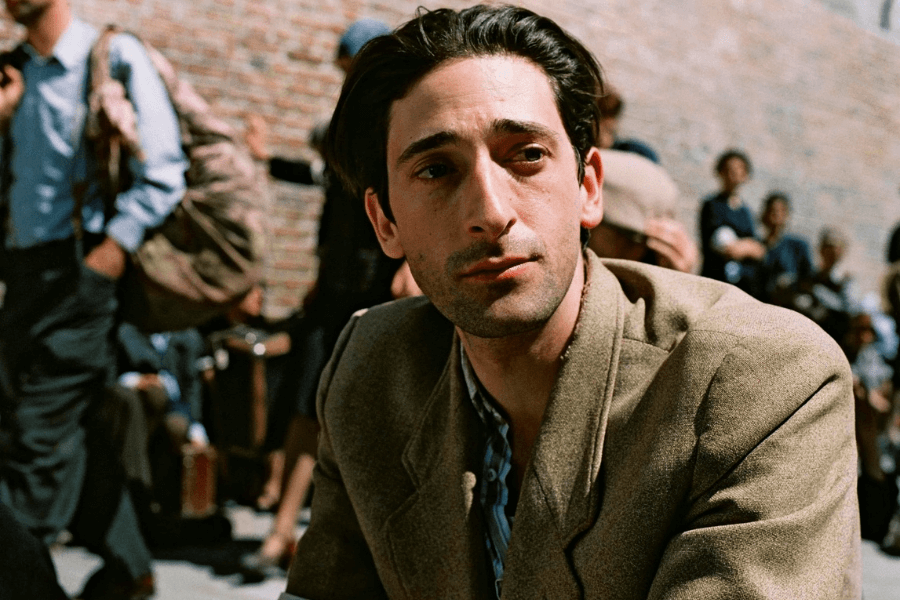 This image shows a man with a contemplative expression, wearing a brown blazer and sitting outdoors under natural light. The background features a crowd of people, some carrying luggage, against a brick wall, suggesting a historical or dramatic setting.