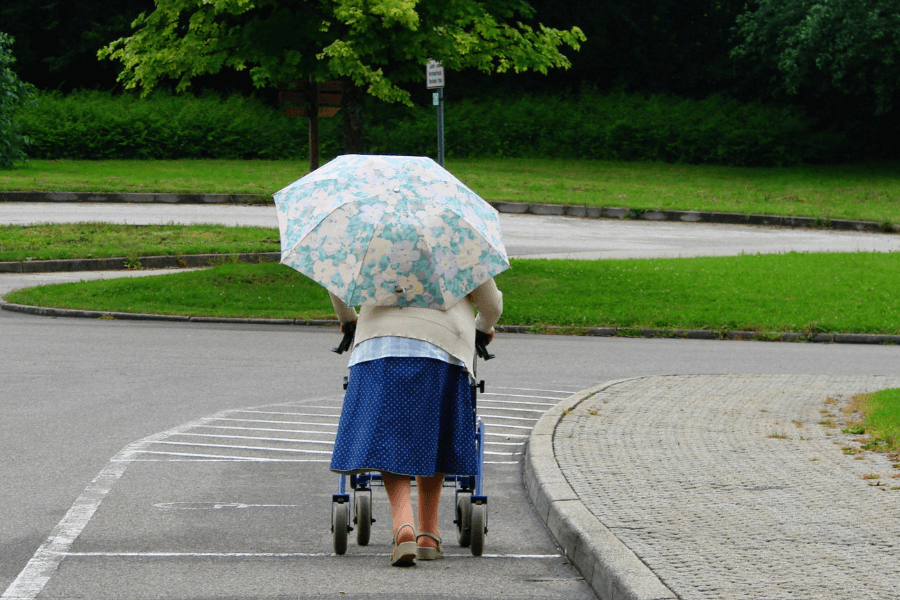 An elderly woman walks outdoors using a walker, holding a floral-patterned umbrella. She is on a paved path surrounded by grass and trees, creating a peaceful park setting.