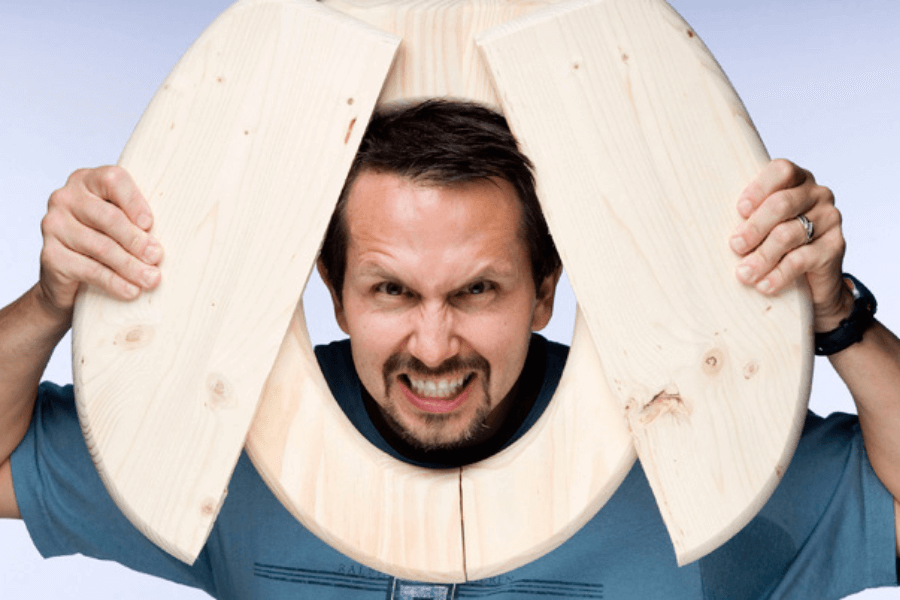 The image shows a man playfully posing with a large wooden object shaped like a toilet seat around his neck. He is making a humorous, exaggerated expression with a scrunched face and clenched teeth, adding a comedic tone to the shot. The clean, light background keeps the focus on his playful and quirky pose.