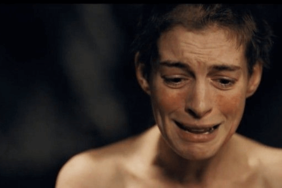 This image depicts a close-up of a woman with a shaved head and tear-streaked face, expressing deep anguish and despair. Her bare shoulders and raw emotion suggest a vulnerable and dramatic moment in a dark setting.