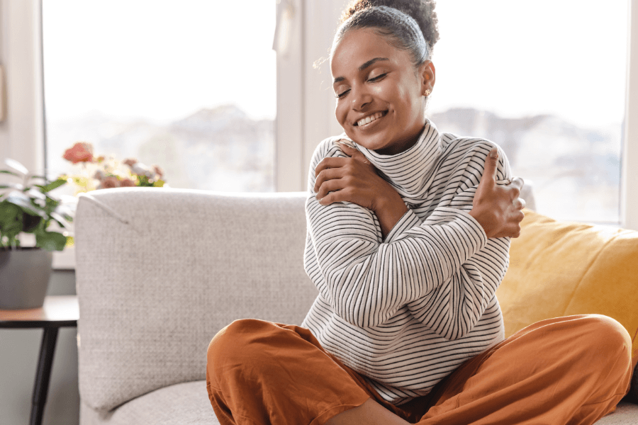 A young woman sits cross-legged on a cozy couch, hugging herself with a joyful expression. She is wearing a striped turtleneck and orange pants, with plants and soft light filling the background.