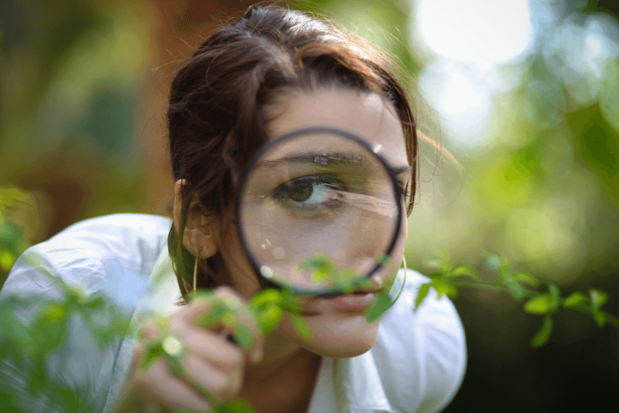 A woman closely examines a plant outdoors using a magnifying glass, her eye enlarged through the lens. The background is lush and green, suggesting a natural or garden setting.